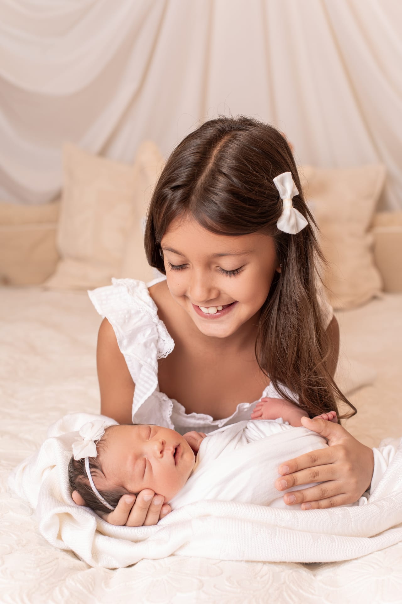 Irmã mais velha sorridente com laço branco no cabelo a olhar carinhosamente para o recém-nascido com tiara enrolado em wrap branco em cama com almofadas creme, Susana Schmitz Fotografia, Porto
