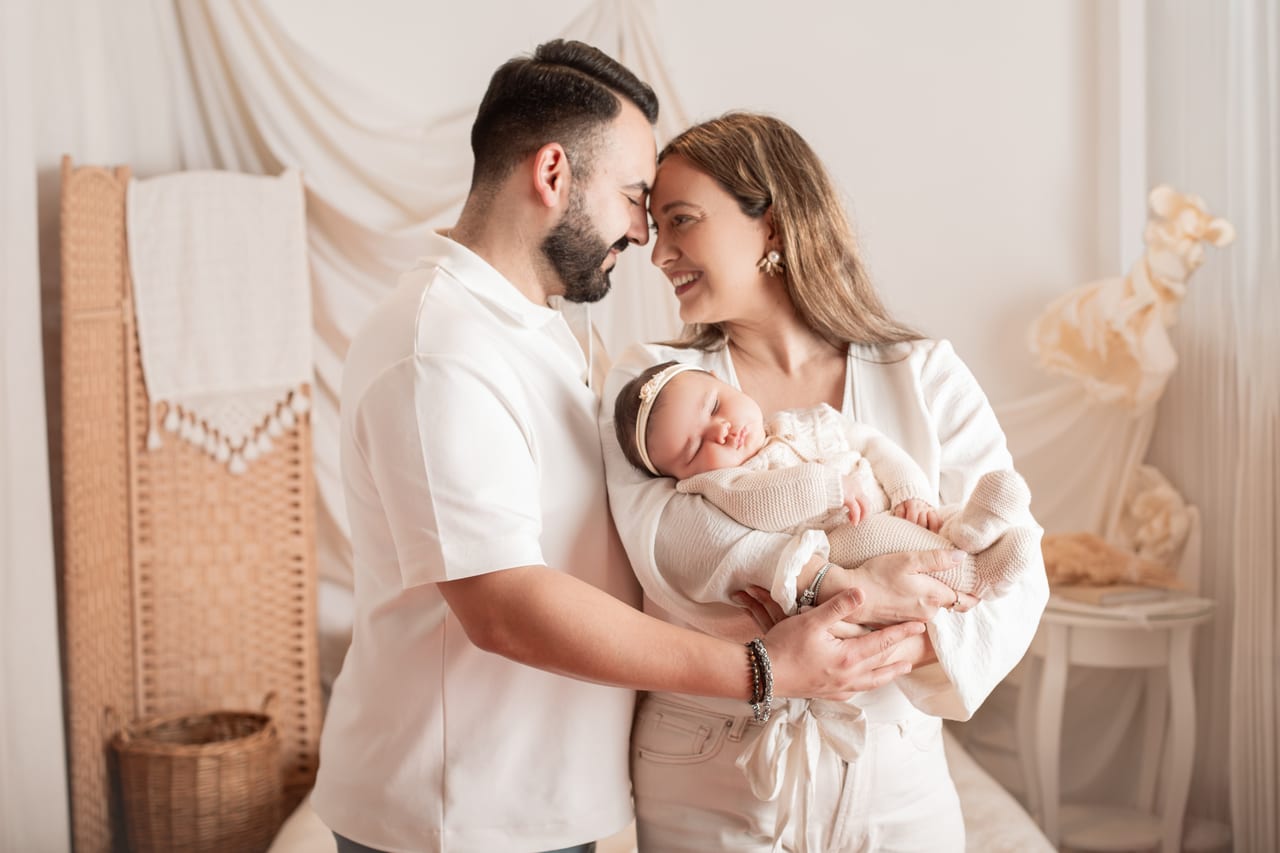Pais sorridentes testa-com-testa em ambiente lifestyle de quarto, mãe a segurar o recém-nascido com tiara enrolado em manta bege, Susana Schmitz Fotografia